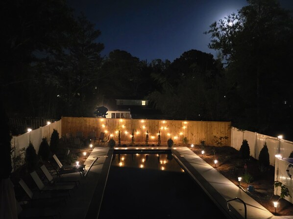 Une piscine dotée d’une terrasse en bois et de chaises longues, entourée d’une clôture en bois agrémentée de guirlandes lumineuses.
