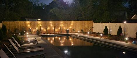 Un área de piscina exterior de noche con sillones de descanso, una cerca de madera y luces colgantes.