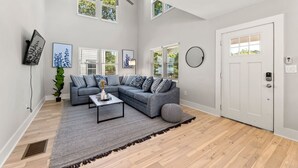 Un salon moderne avec un canapé d’angle, une table basse et un ventilateur de plafond. De nombreuses fenêtres laissent entrer la lumière naturelle et offrent une vue sur l’extérieur.
