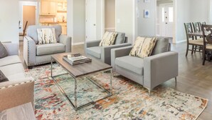 Un salon spacieux agrémenté d’un lustre moderne, de canapés gris ornés de coussins à motifs, d’une table basse et d’un tapis coloré.