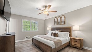 A bedroom with a wooden bed, two bedside tables with lamps, a ceiling fan, a large window, and a TV mounted on the wall. - Beautiful Newly-renovated Home in Indian Hills (Marietta)