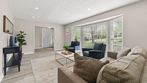 A spacious living room with a large couch, a coffee table, and a side table with decorative items. There are two windows with blinds, a door leading to another room, and a ceiling with recessed lighting. - Beautiful Newly-renovated Home in Indian Hills (Marietta)