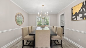 A dining room with a long table, six chairs, a chandelier, a window with blinds, and a painting on the wall. - Beautiful Newly-renovated Home in Indian Hills (Marietta)