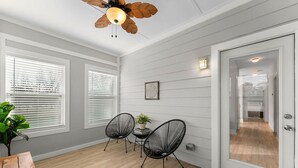 Ett rum med vitmålade panelväggar, ett trägolv, en takfläkt, två svarta stolar, ett litet bord med en växt och en dörr som leder till ett annat rum.