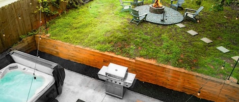 A backyard with a hot tub, a grill, and a fire pit surrounded by chairs.