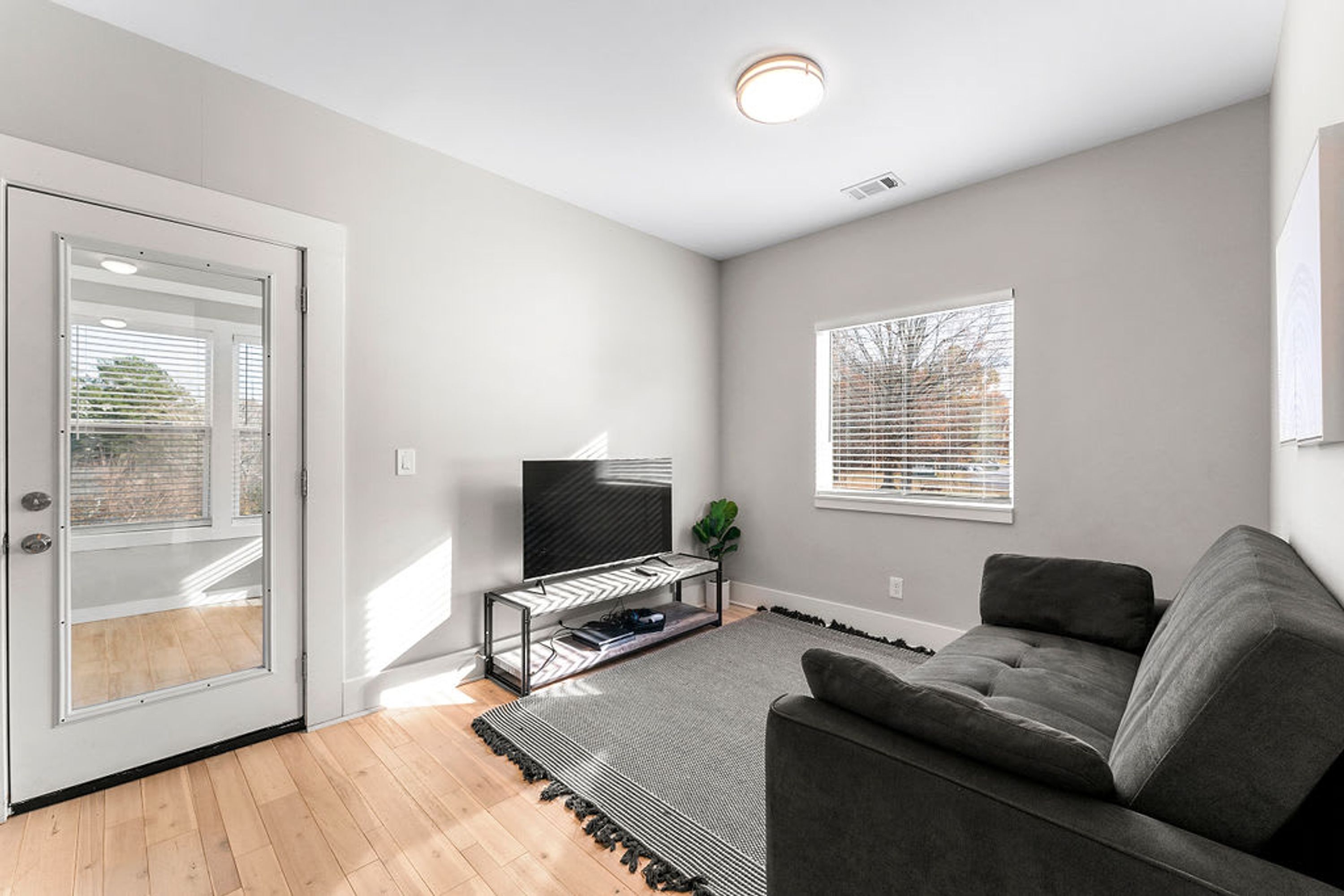 A modern living room with a grey sofa, a TV on a stand, a glass door, and a window with blinds.