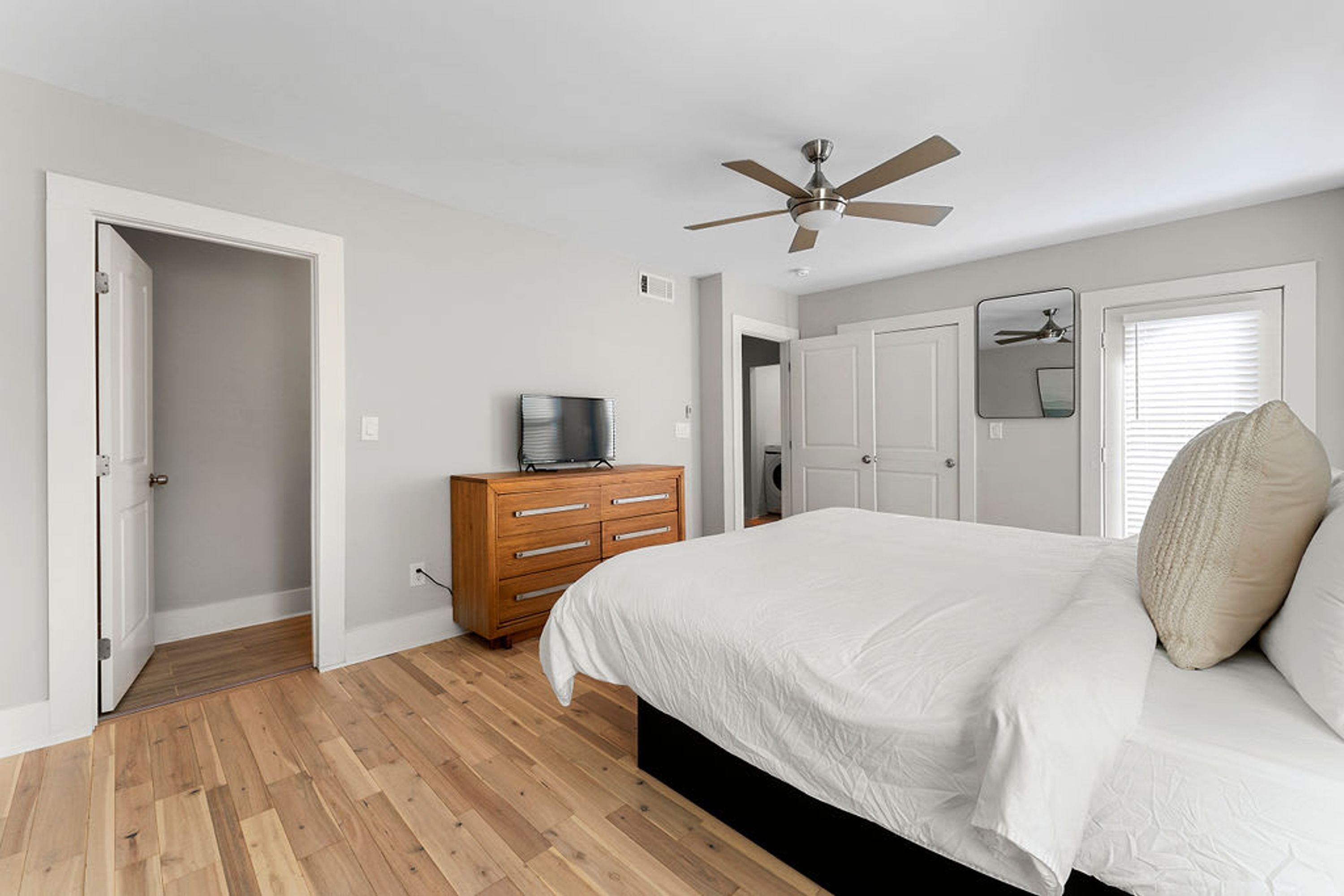 Een slaapkamer met een houten vloer, een bed met witte beddengoed, een houten dressoir, een plafondventilator en een deur naar een andere kamer.