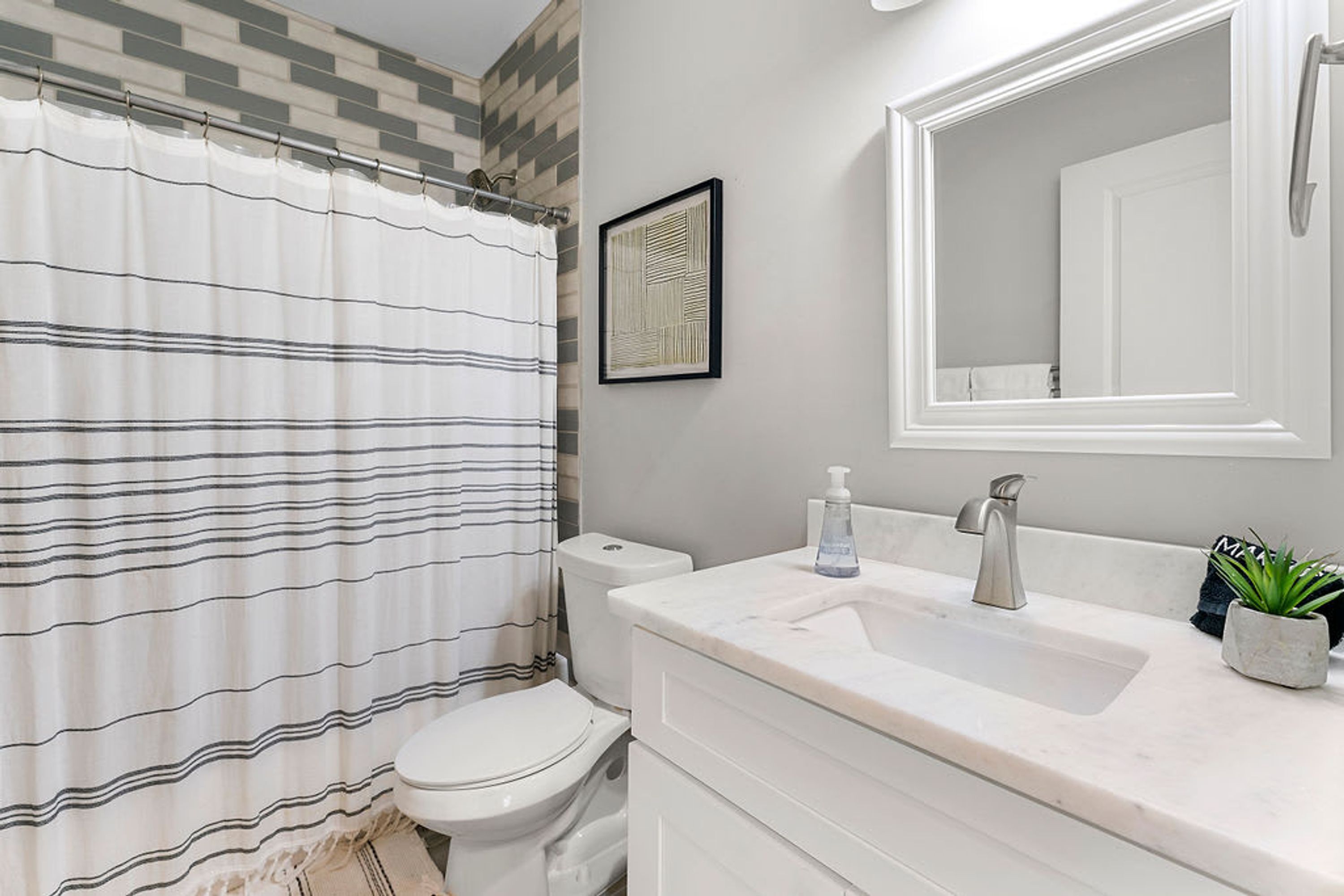 A bathroom with a striped shower curtain, a toilet, a sink with a marble countertop, a mirror, and a framed artwork on the wall.