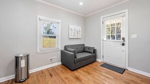 A room with a grey sofa, a white door, and a window with blinds. - Private 1BR Cottage In Downtown Historic Norcross (Norcross)
