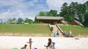A beach scene with a sandy area, a water body, and a building with a roof in the background. - Private Resort - Lake Lanier Paradise (Gainesville)