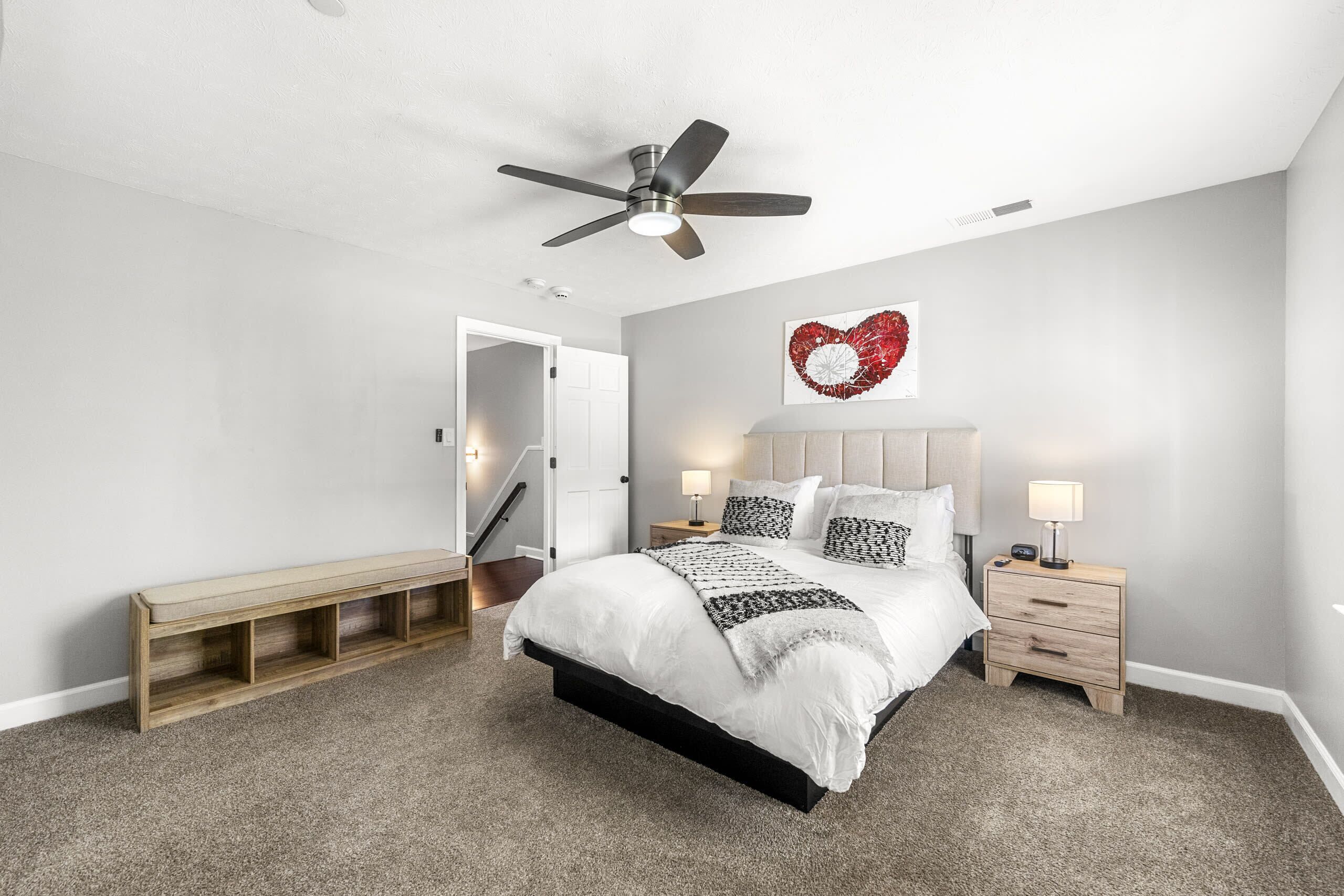 Un dormitorio con cama, mesitas de noche, un banco, ventilador de techo y una obra de arte con forma de corazón en la pared.