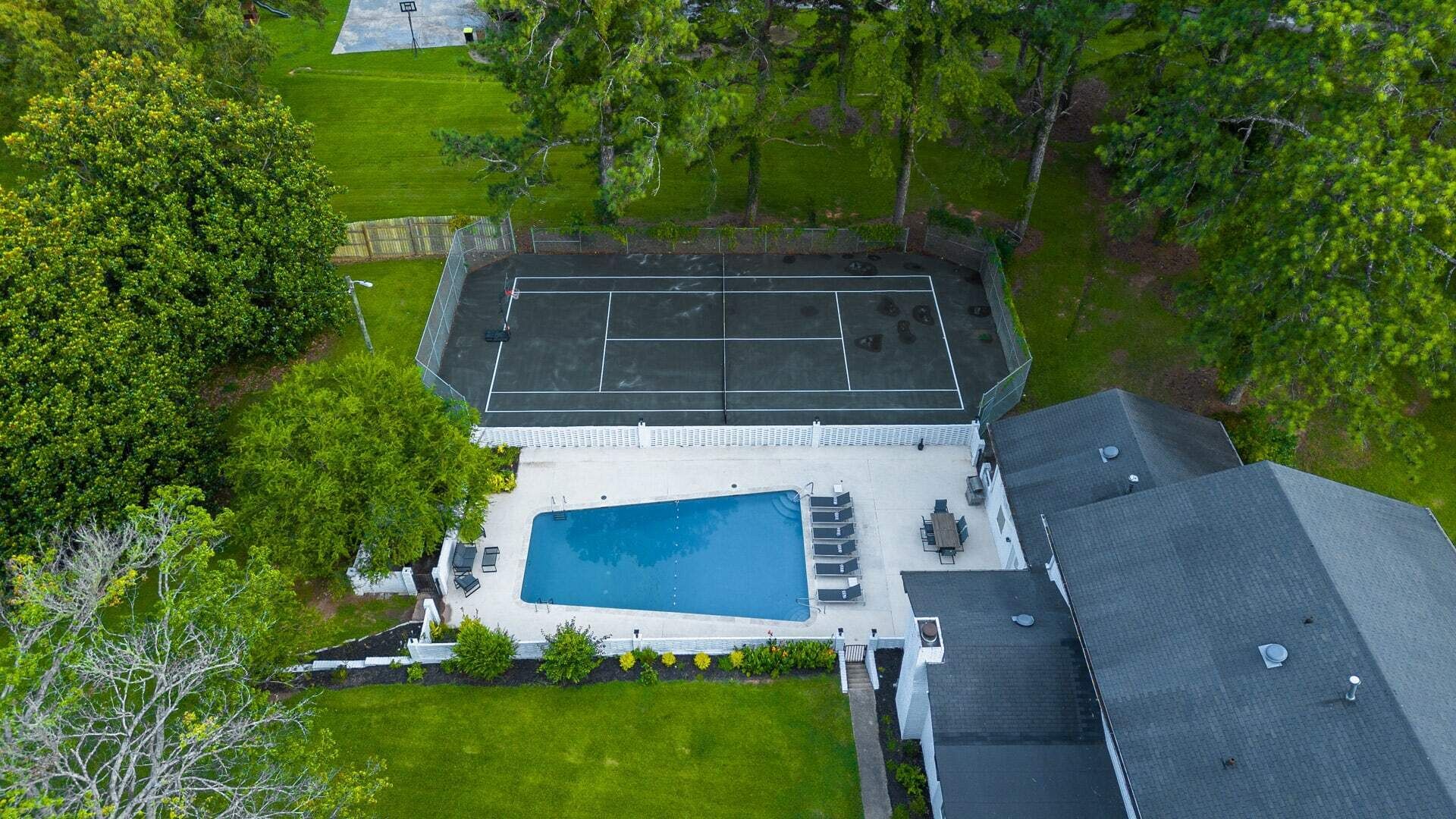 An aerial view of a residential property with a swimming pool, a tennis court, and a fenced yard.