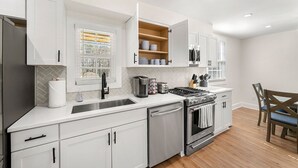A modern kitchen with white cabinetry, stainless steel appliances, and a window with blinds. - Beautifully Modern Cottage in Historic Norcross (Norcross)