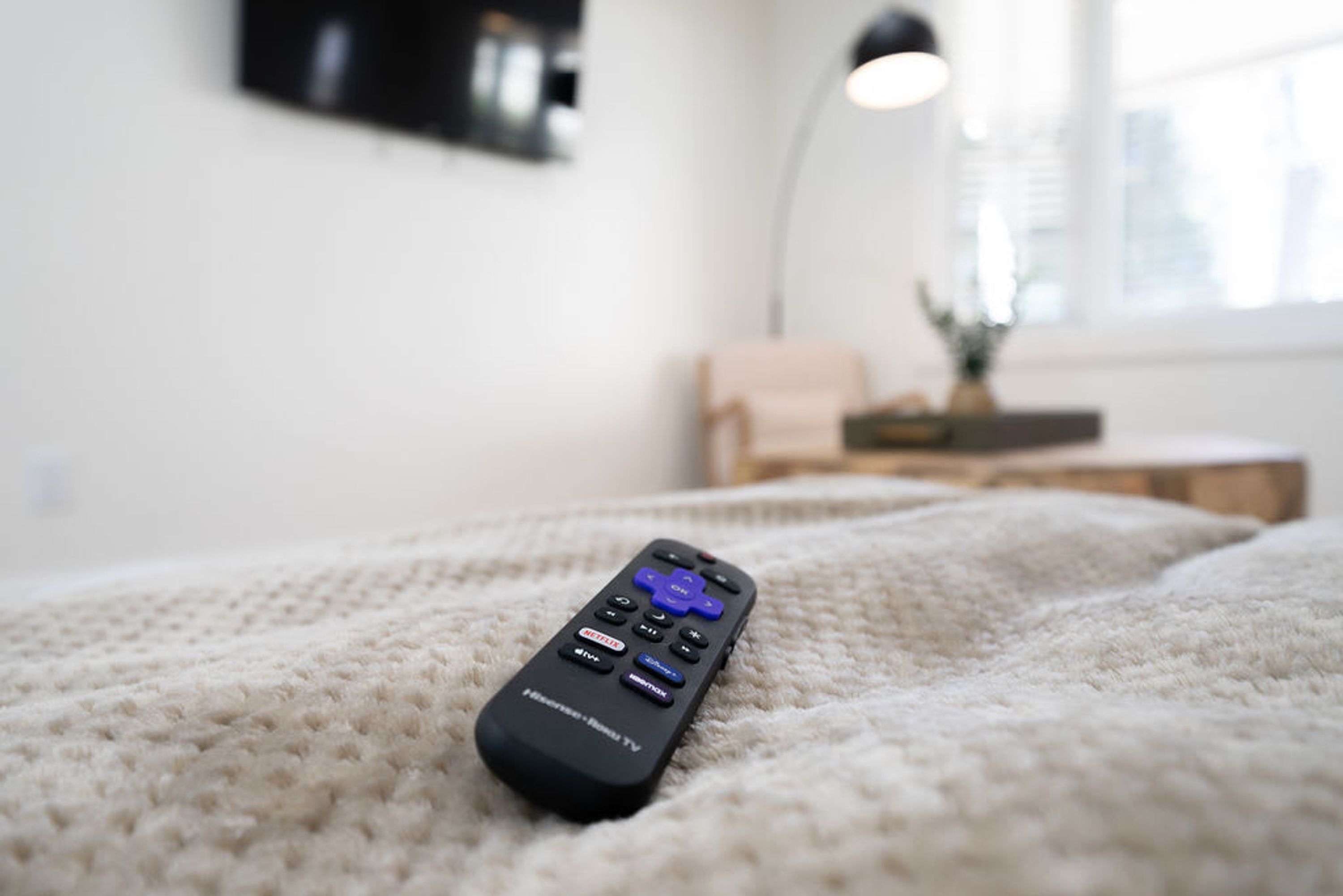 A remote control on a textured white blanket in a room with a lamp and a plant on a side table.