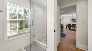 A modern bathroom with a glass shower enclosure, a white tiled shower area, and a window with blinds. - Beautifully Modern Cottage in Historic Norcross (Norcross)