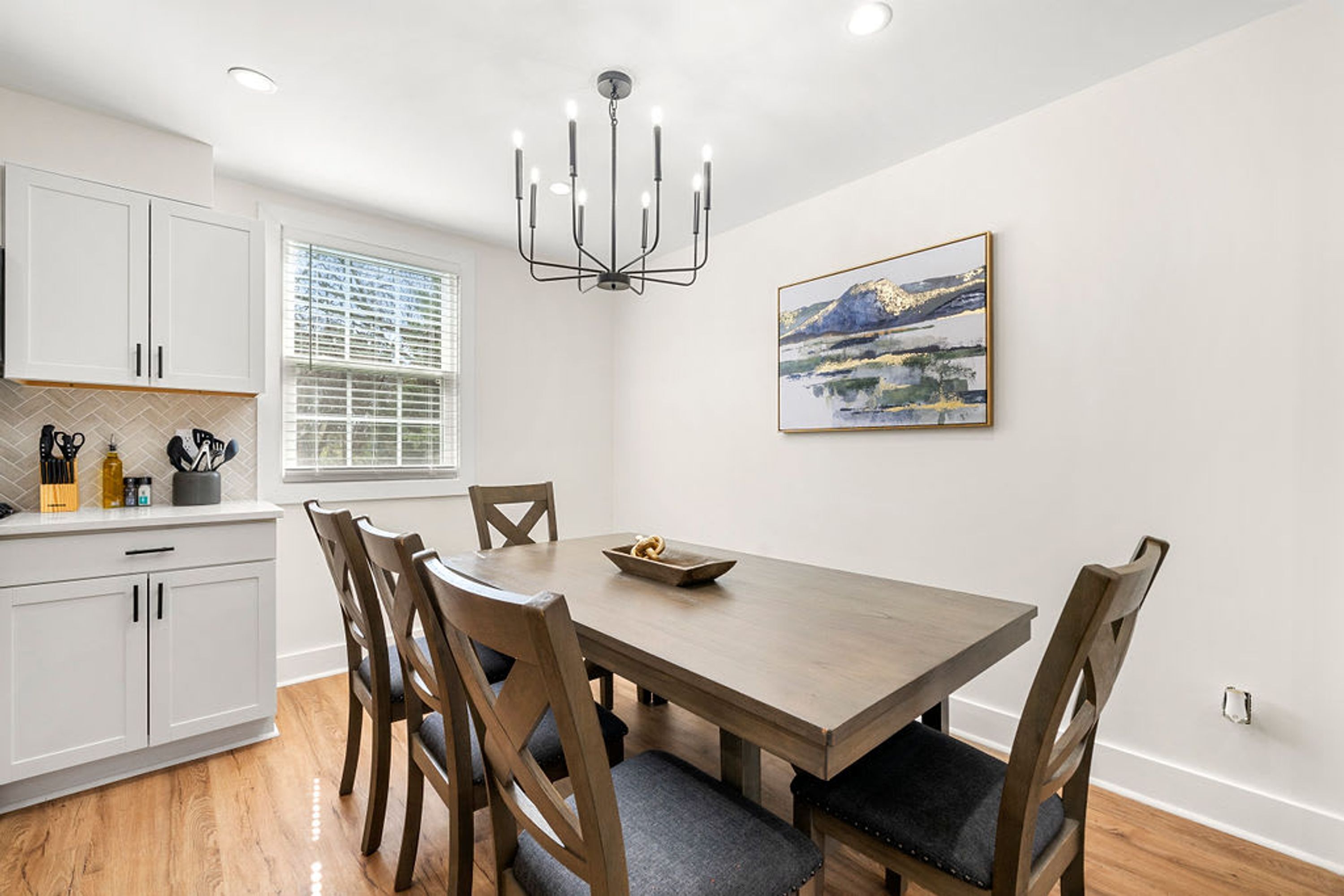 A dining area with a wooden table and chairs, a chandelier, a window with blinds, and a painting on the wall.