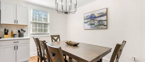 A dining area with a wooden table and chairs, a chandelier, a window with blinds, and a painting on the wall.