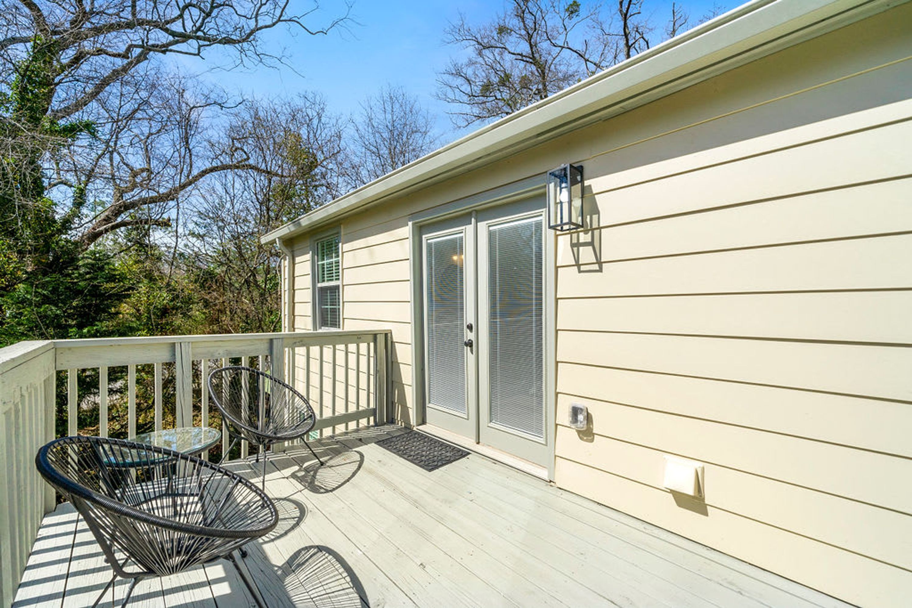 A covered outdoor deck with two metal chairs and a small table, a sliding glass door, and a light fixture on the side of a beige building.