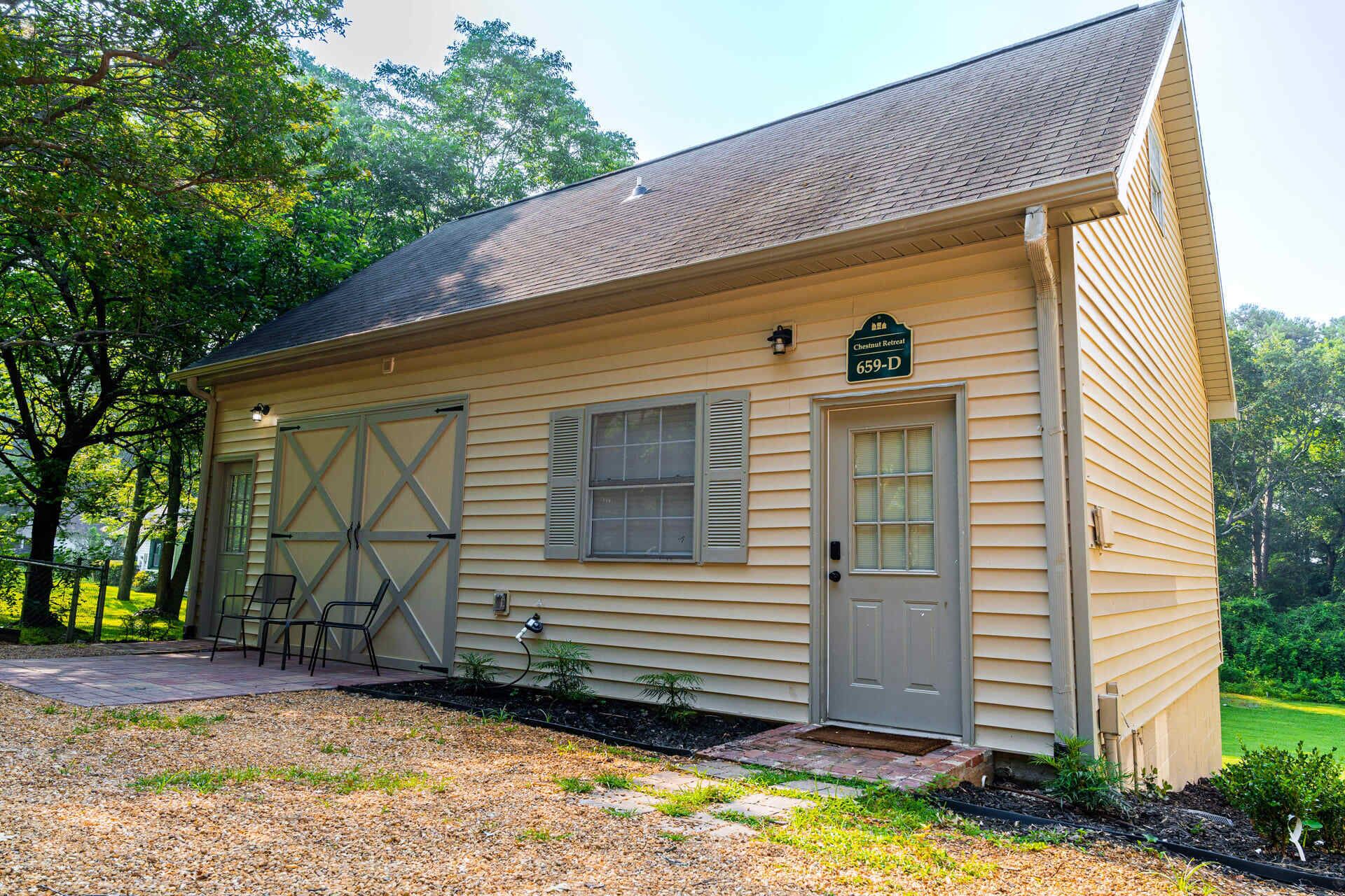 A single-story building with a gabled roof, a garage door, and a small porch with chairs.