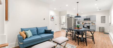 A modern living room with a blue sofa, a dining table with chairs, and a kitchen area in the background.