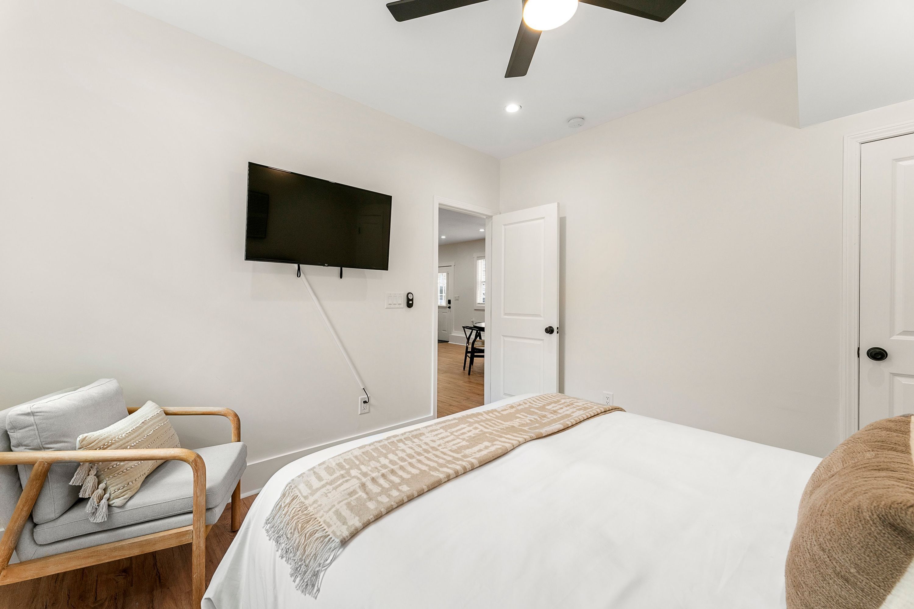A bedroom with a bed, a TV mounted on the wall, a ceiling fan, and a door leading to another room.