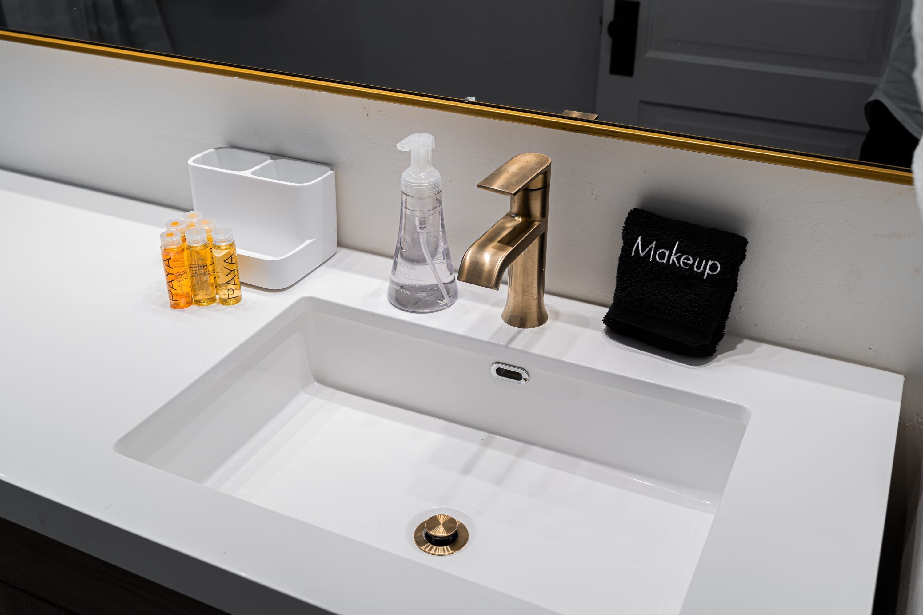 Un lavamanos blanco con grifo dorado, una toalla para maquillaje, un dispensador de jabón y un recipiente con artículos de tocador.