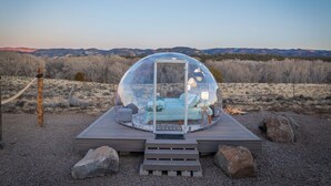 A transparent geodesic dome structure with a bed and seating area, surrounded by a wooden deck and gravel, set against a mountainous backdrop. - Brycegate Retreat, Cabin, Star Dome Bedroom, Fire Pit, Hammocks, Views, Pets, EV (Hatch)