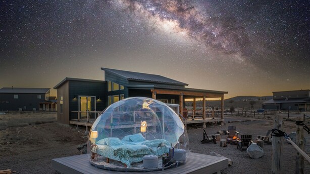 A geodesic dome structure with a bed inside, set on a wooden platform, surrounded by a residential area with houses and a clear night sky. - Brycegate Retreat, Cabin, Star Dome Bedroom, Fire Pit, Hammocks, Views, Pets, EV (Hatch)