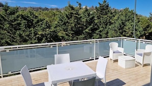 Un balcón con barandilla de cristal, mobiliario de mimbre blanco y vista a una ladera boscosa.