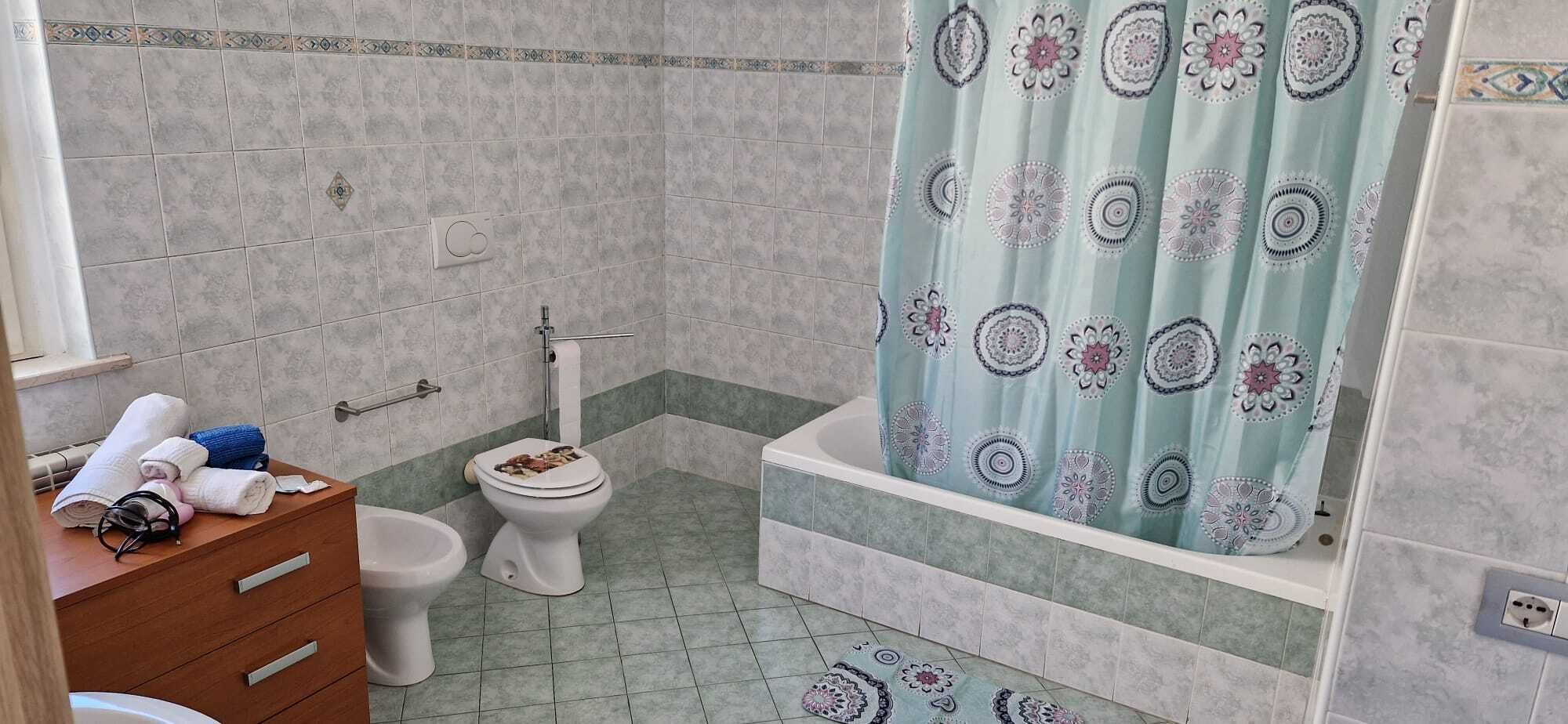 A bathroom with a toilet, bidet, and bathtub. There is a towel rack with towels and a small wooden cabinet.