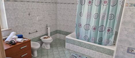 A bathroom with a toilet, bidet, and bathtub. There is a towel rack with towels and a small wooden cabinet.