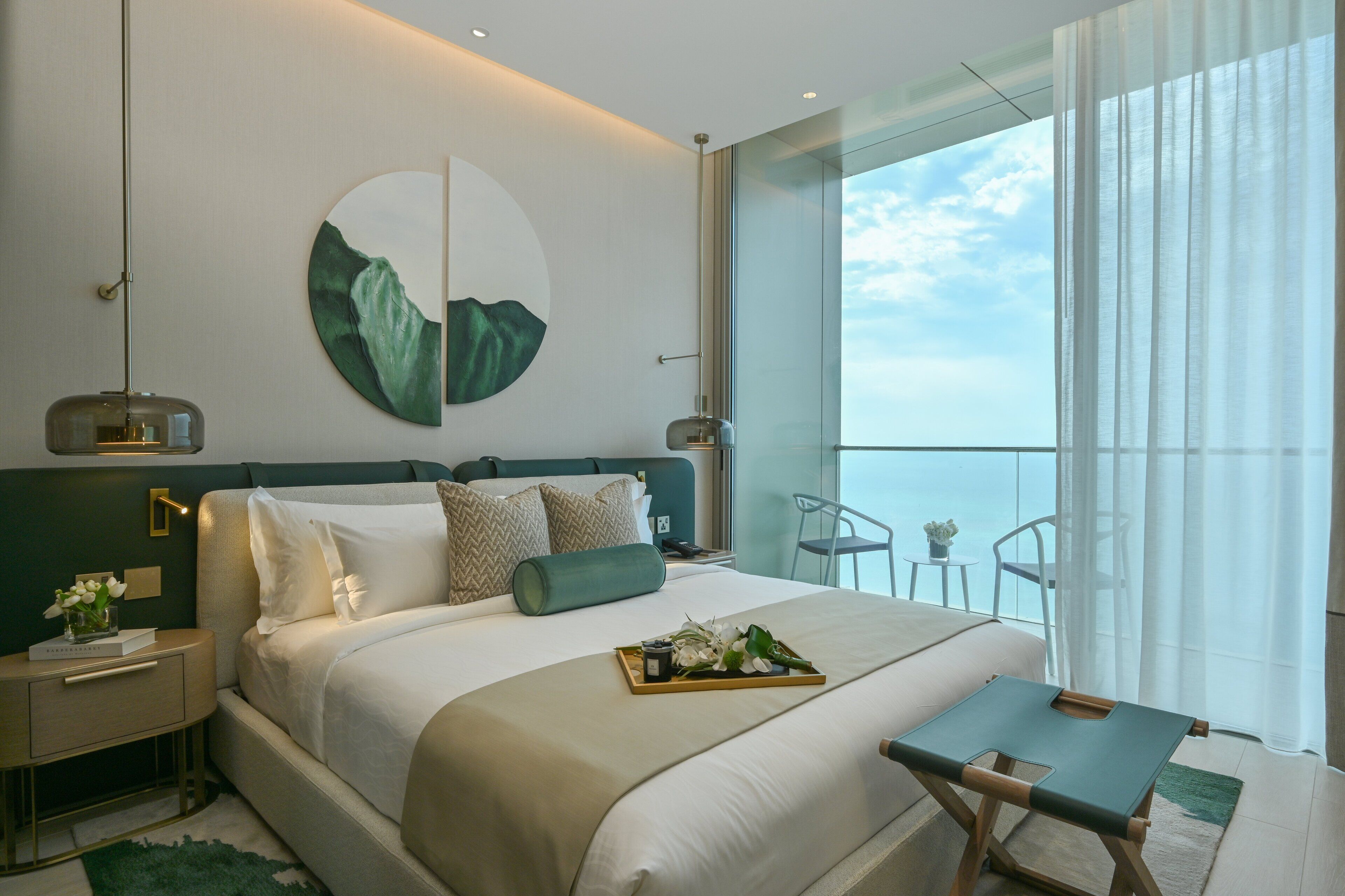 Una habitación de hotel moderna con una cama grande, una mesita de noche con lámpara, vista al mar desde un balcón y una obra de arte circular en la pared.