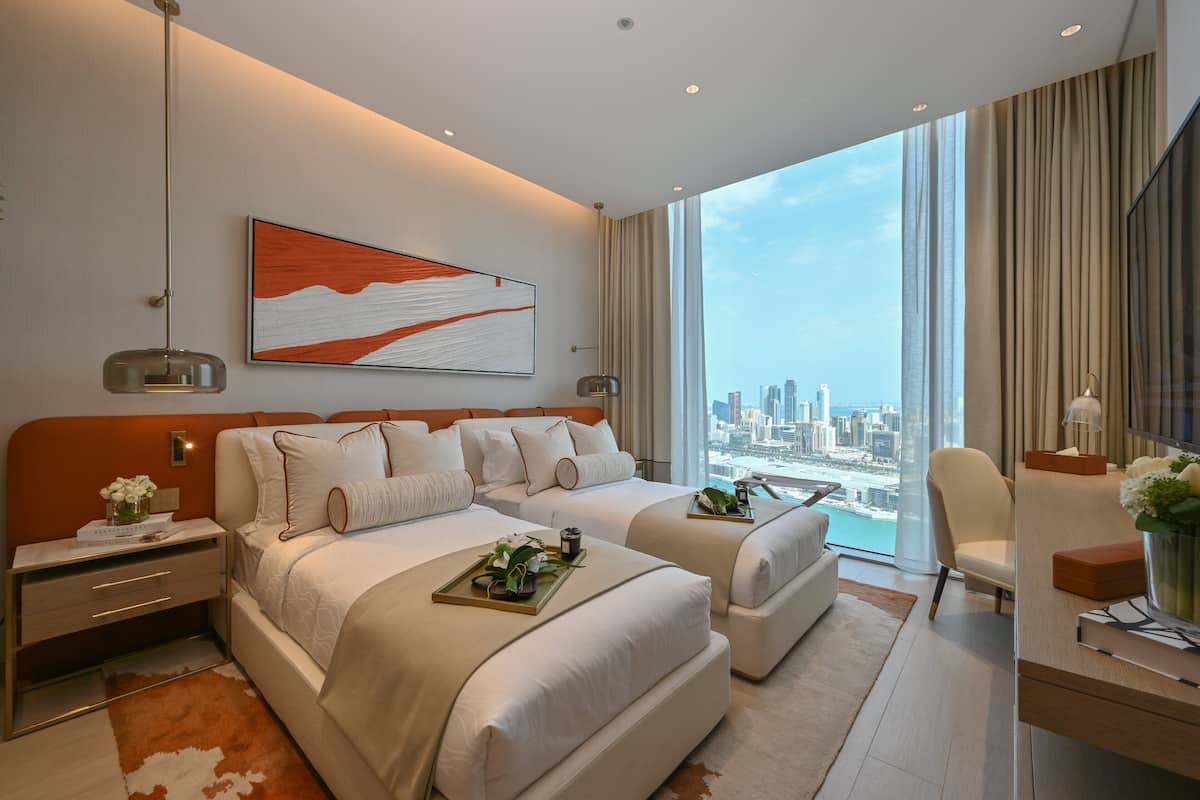 A modern hotel room with two beds, a city view, a large abstract painting, and a desk with a lamp.