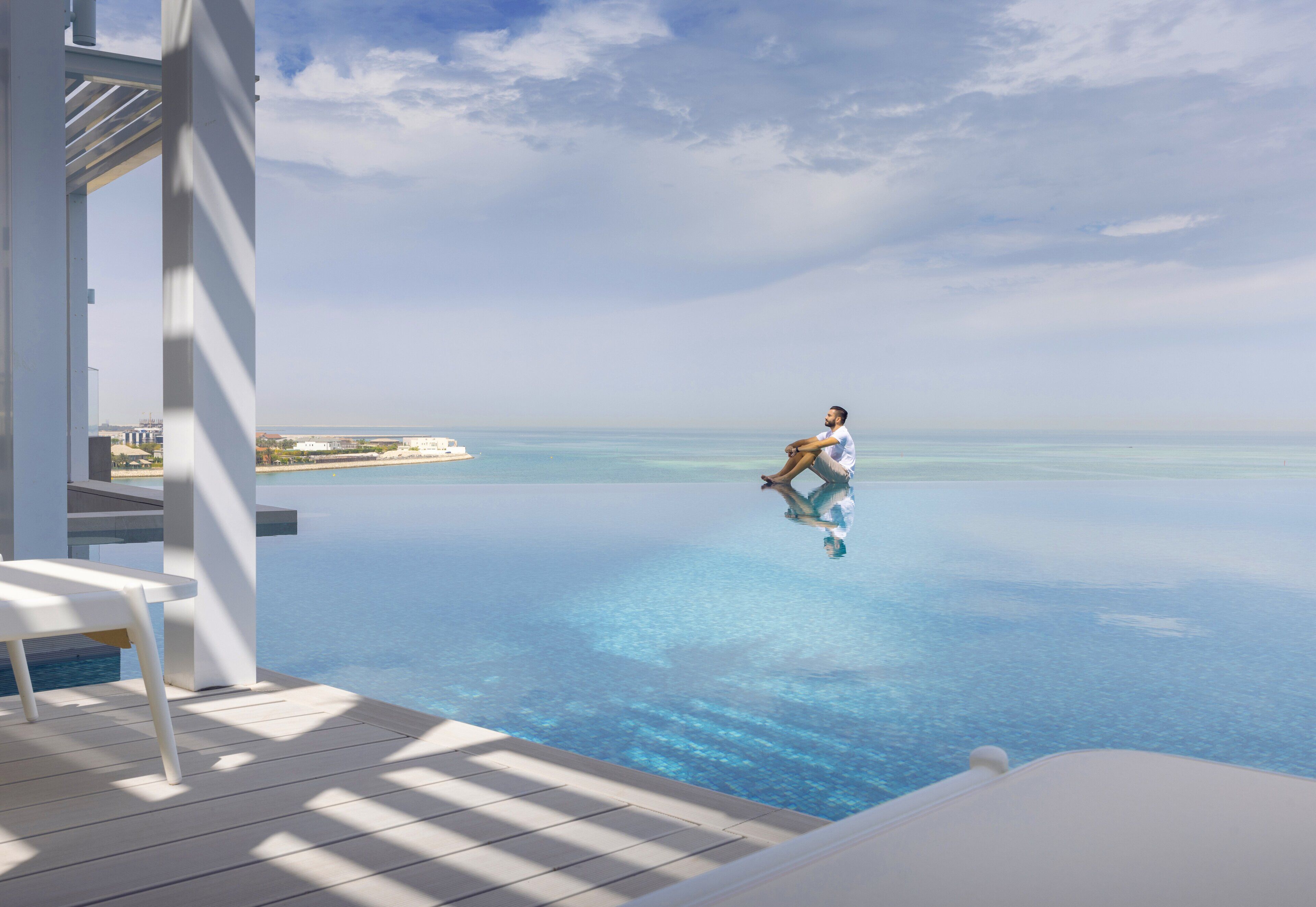 Un hombre descansando en una piscina infinita con vista despejada al océano y edificios lejanos.