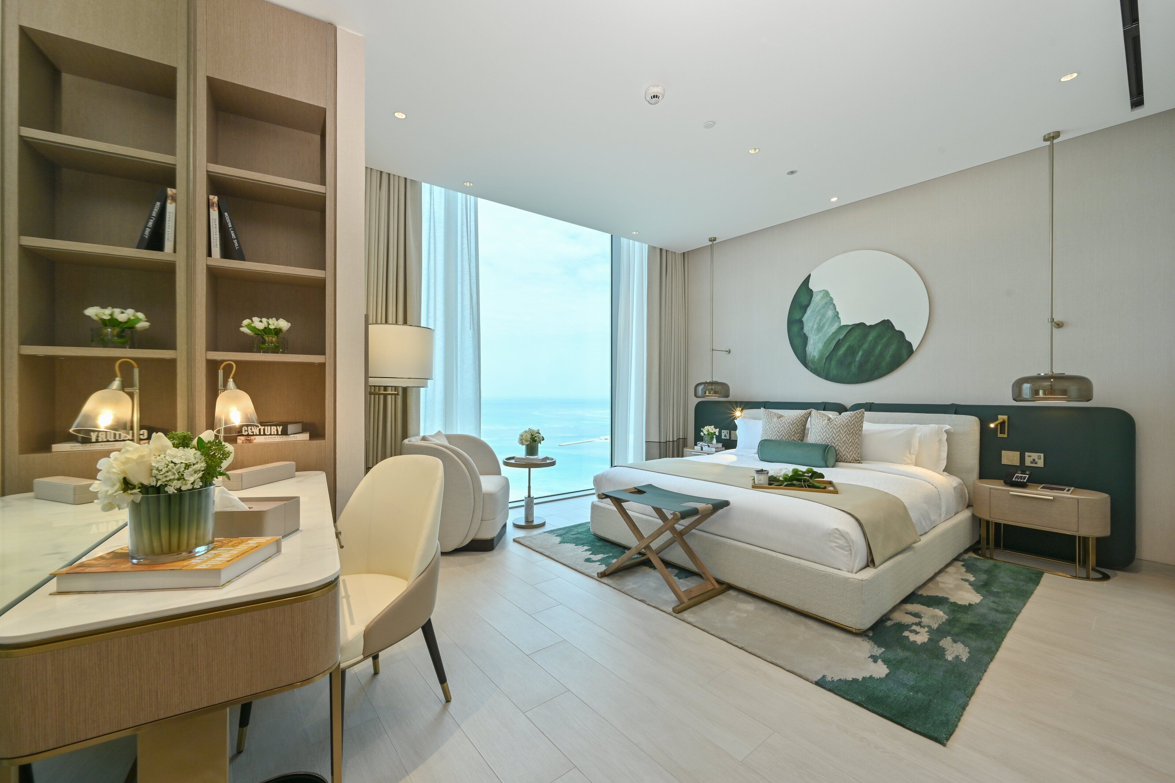 Una habitación de hotel moderna con una cama grande, un escritorio con silla, una estantería, un espejo redondo y vista al mar.