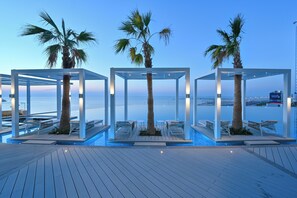 Zona de piscina con tres pérgolas blancas, cada una con una palmera y asientos.