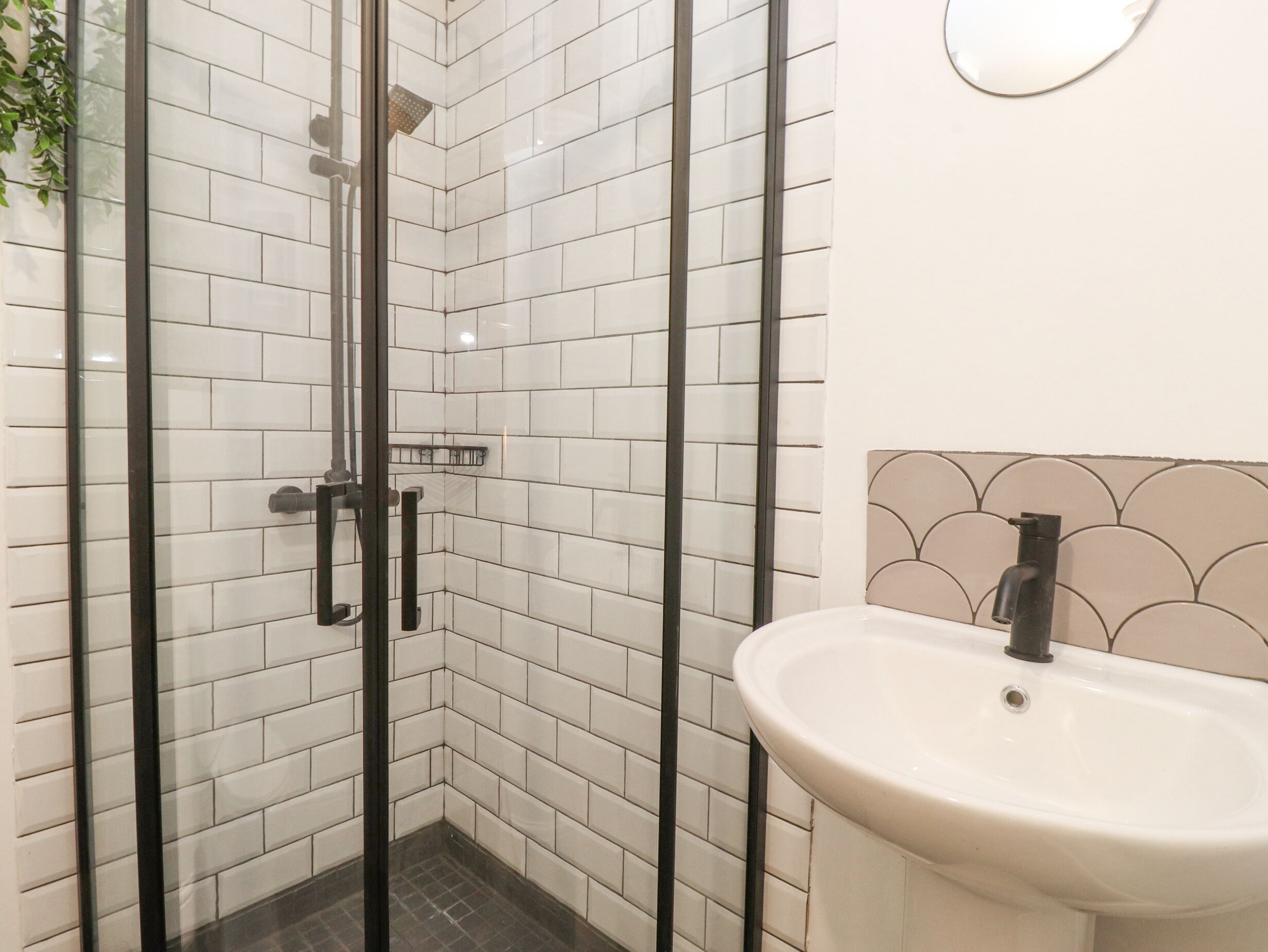 Een badkamer met witte betegelde wanden, een glazen douchecabine, een zwart metalen frame, een wit wastafelmeubel en een beige spatwand, en een spiegel aan de muur.