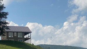 A modern house with a metal roof, situated on a grassy hill with a clear sky and clouds.
