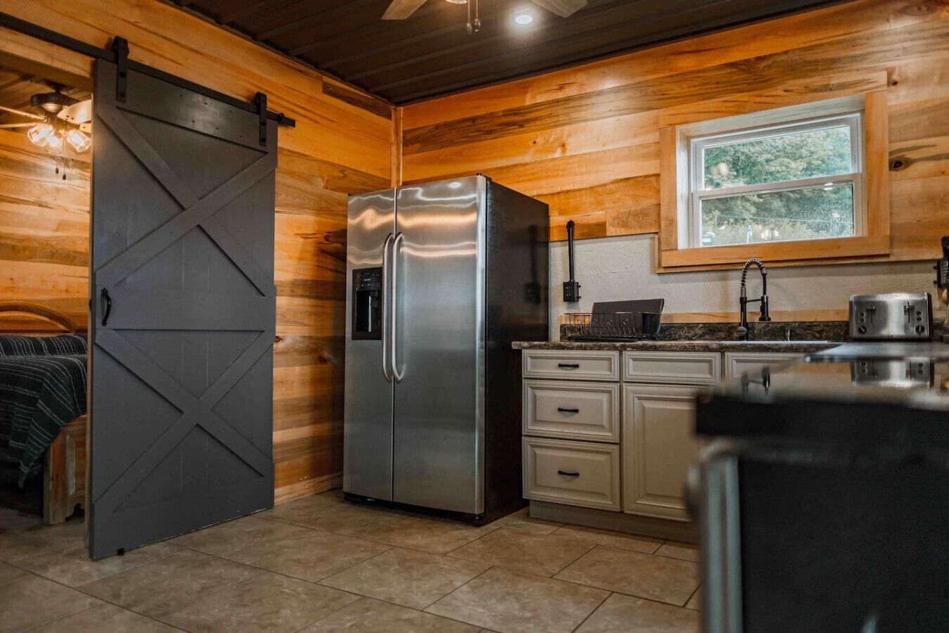 A kitchen with a stainless steel refrigerator, wooden cabinets, a window, and a sliding barn door.