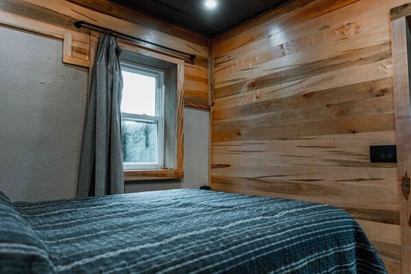 Une pièce aux murs en bois, une fenêtre avec des rideaux et un lit recouvert d’une couverture à rayures.