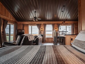 Une chambre avec un grand lit, un bureau et une chaise. Elle est dotée de fenêtres avec des rideaux, d’un ventilateur de plafond et offre une vue sur l’extérieur.