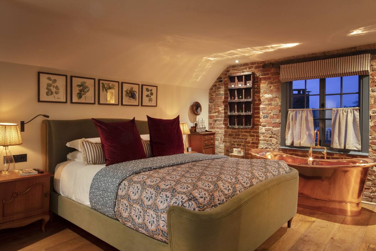 A bedroom with a large bed, a brick wall, a window with curtains, and a wooden floor.