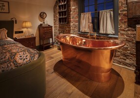 Una habitación con una bañera de cobre grande, muebles de madera, una pared de ladrillo y una ventana con cortinas.