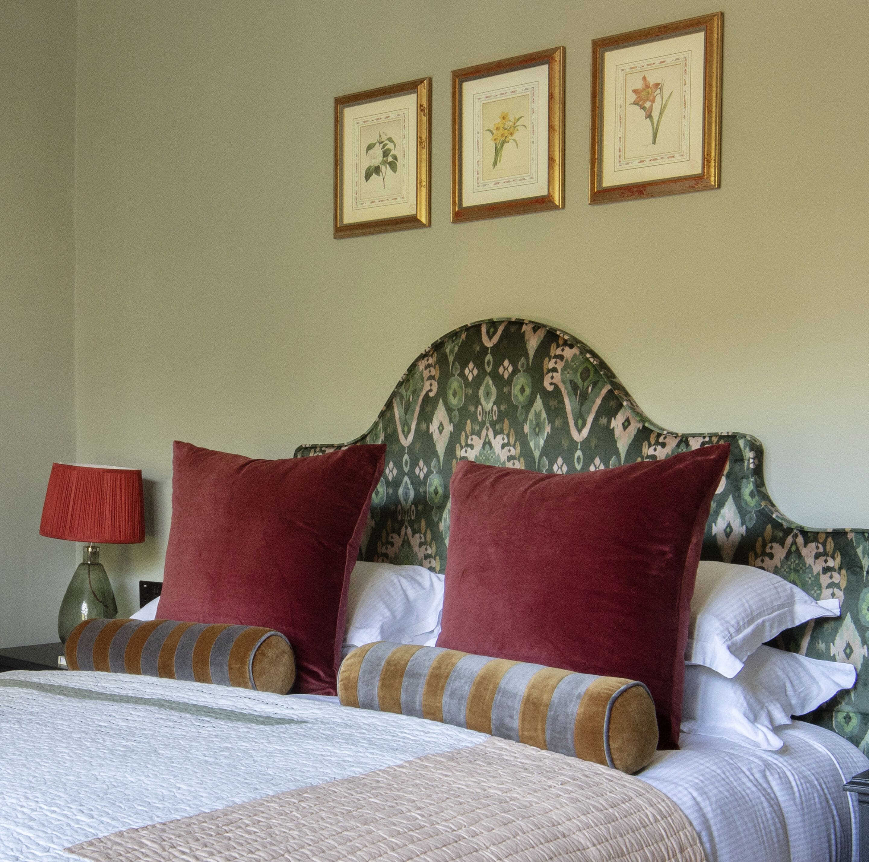 A neatly made bed with red and striped pillows, a green patterned headboard, and three framed botanical prints on the wall.
