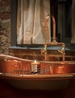 Un lavamanos de cobre con una vela en un candelabro de vidrio, una rejilla metálica y una ventana con cortinas al fondo.