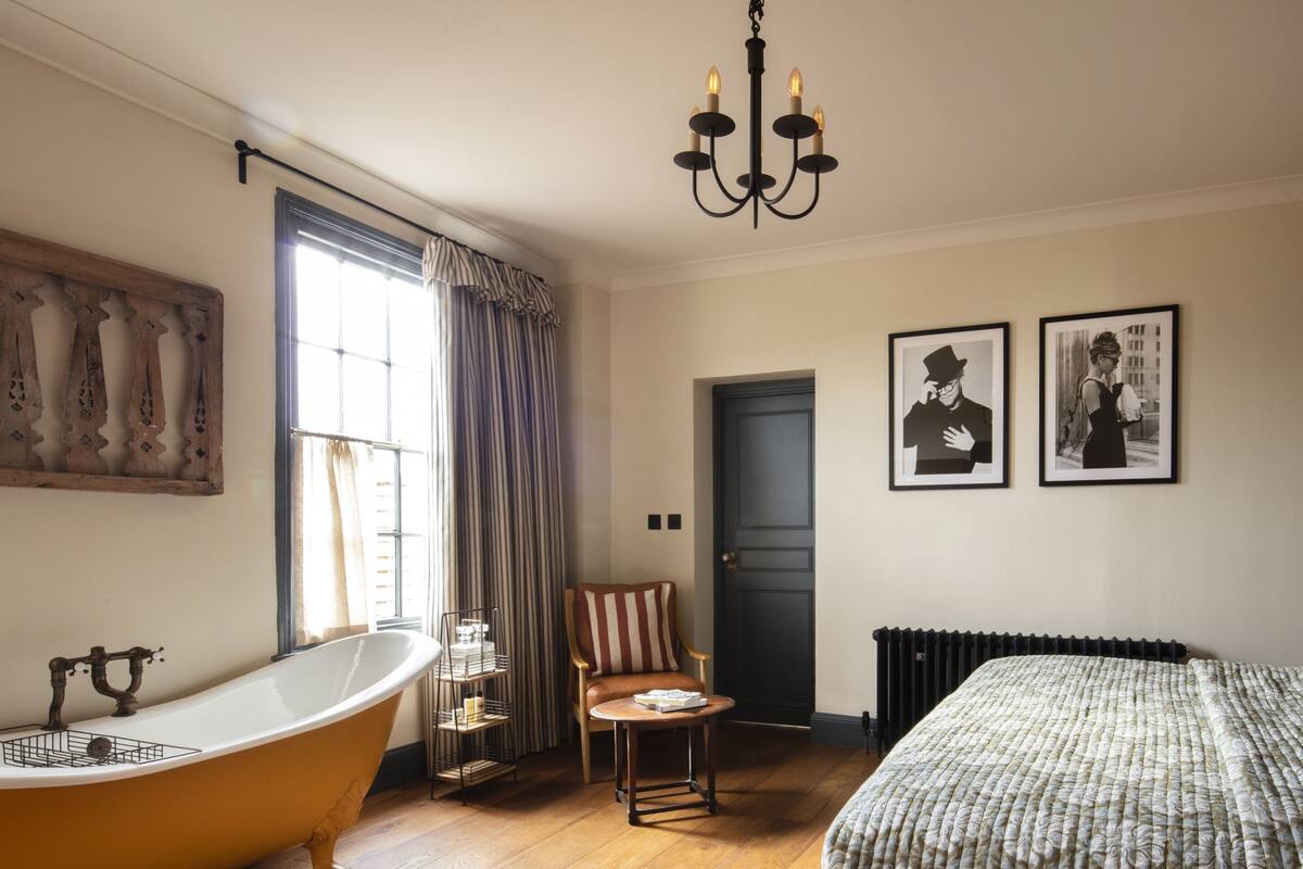 A room with a clawfoot bathtub, a wooden floor, a bed with a patterned duvet, a chair with striped cushions, a chandelier, and two framed pictures on the wall.