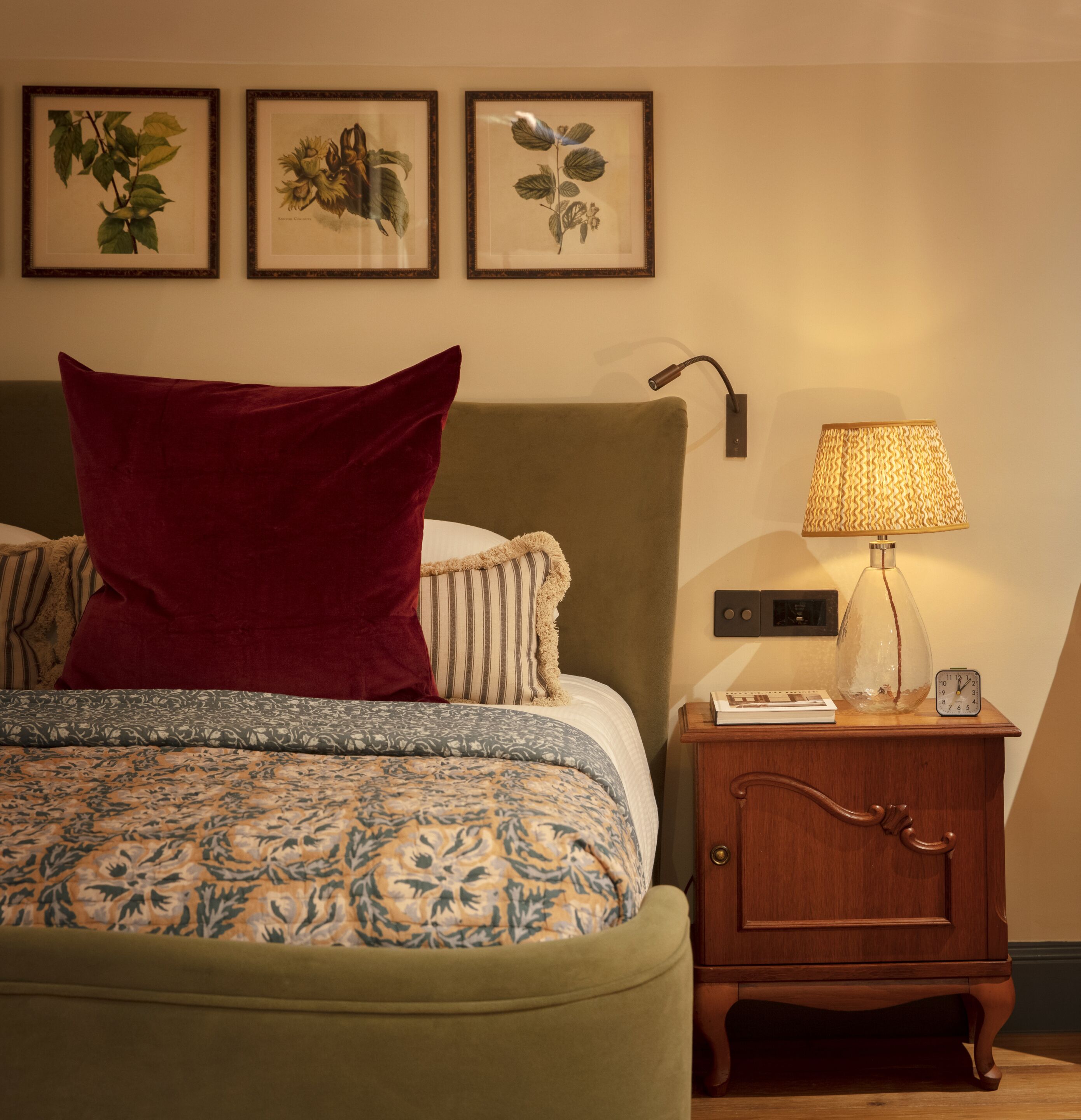 A neatly arranged bedroom with a bed, pillows, a bedside lamp, and framed botanical prints on the wall.
