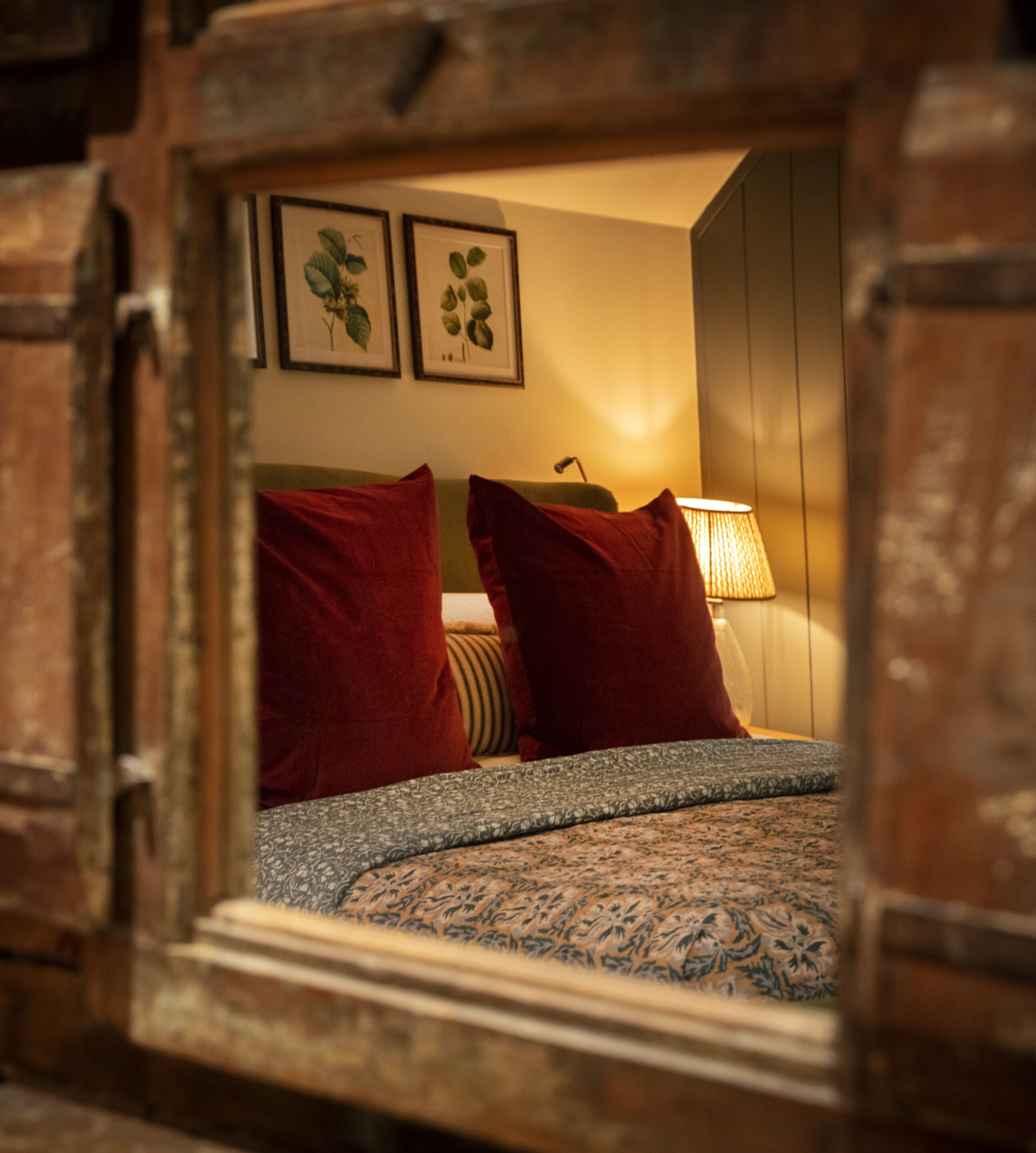 A cozy bedroom with a bed, two red pillows, a patterned bedspread, a lamp, and framed botanical prints on the wall.