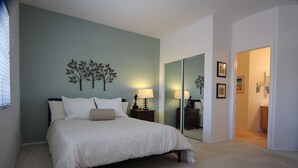 Une chambre à coucher comprenant un lit, une table de chevet, une lampe, un miroir et une porte donnant sur une autre pièce.