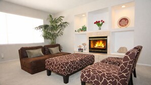 Un salon avec une cheminée, un canapé, deux poufs, une grande plante et des objets de décoration.